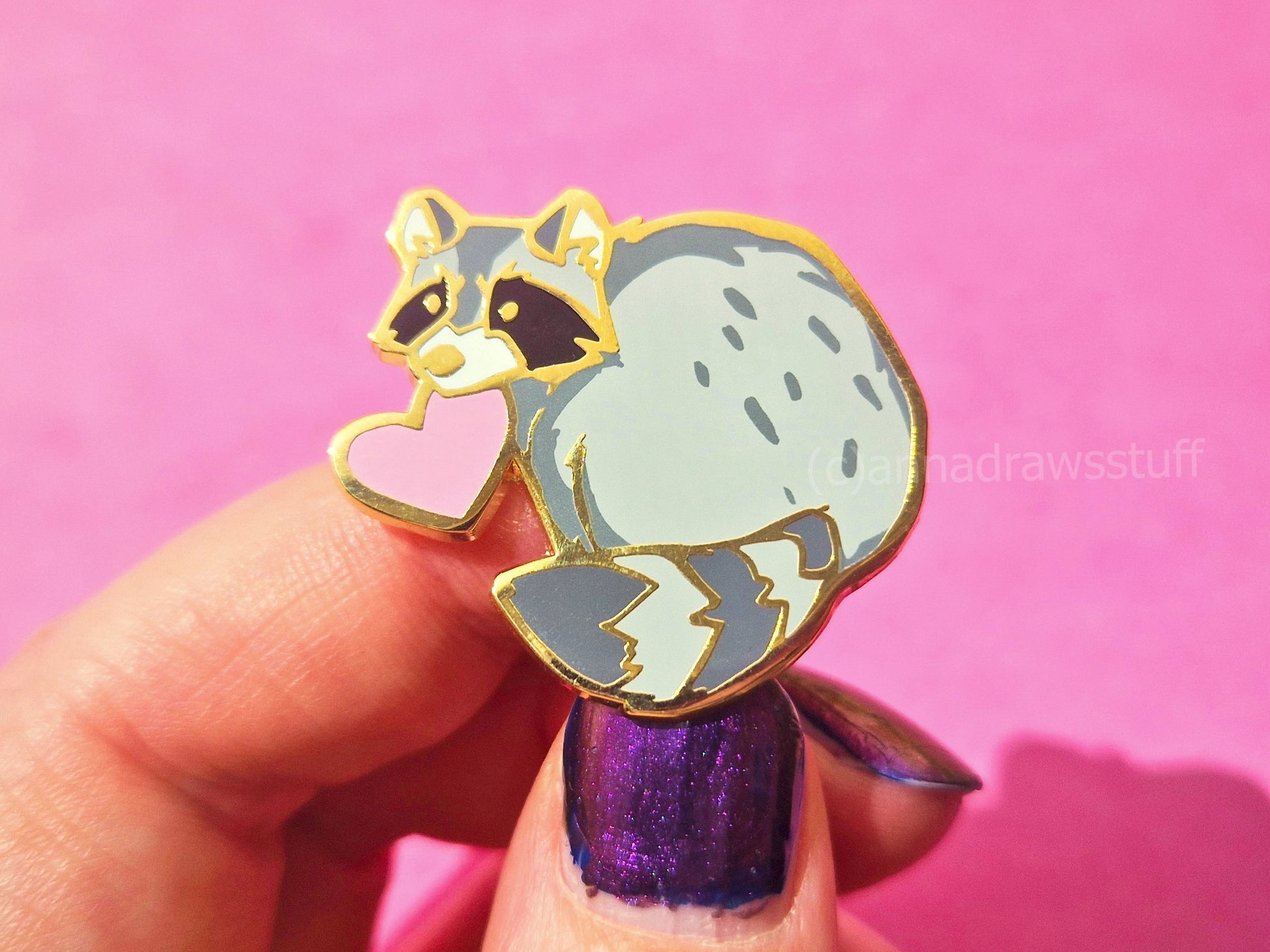 A hand with purple glitter nail polish holds a raccoon enamel pin against a solid pink background. The pin features a light gray raccoon with black facial markings, a pink heart, and gold metal outlines.