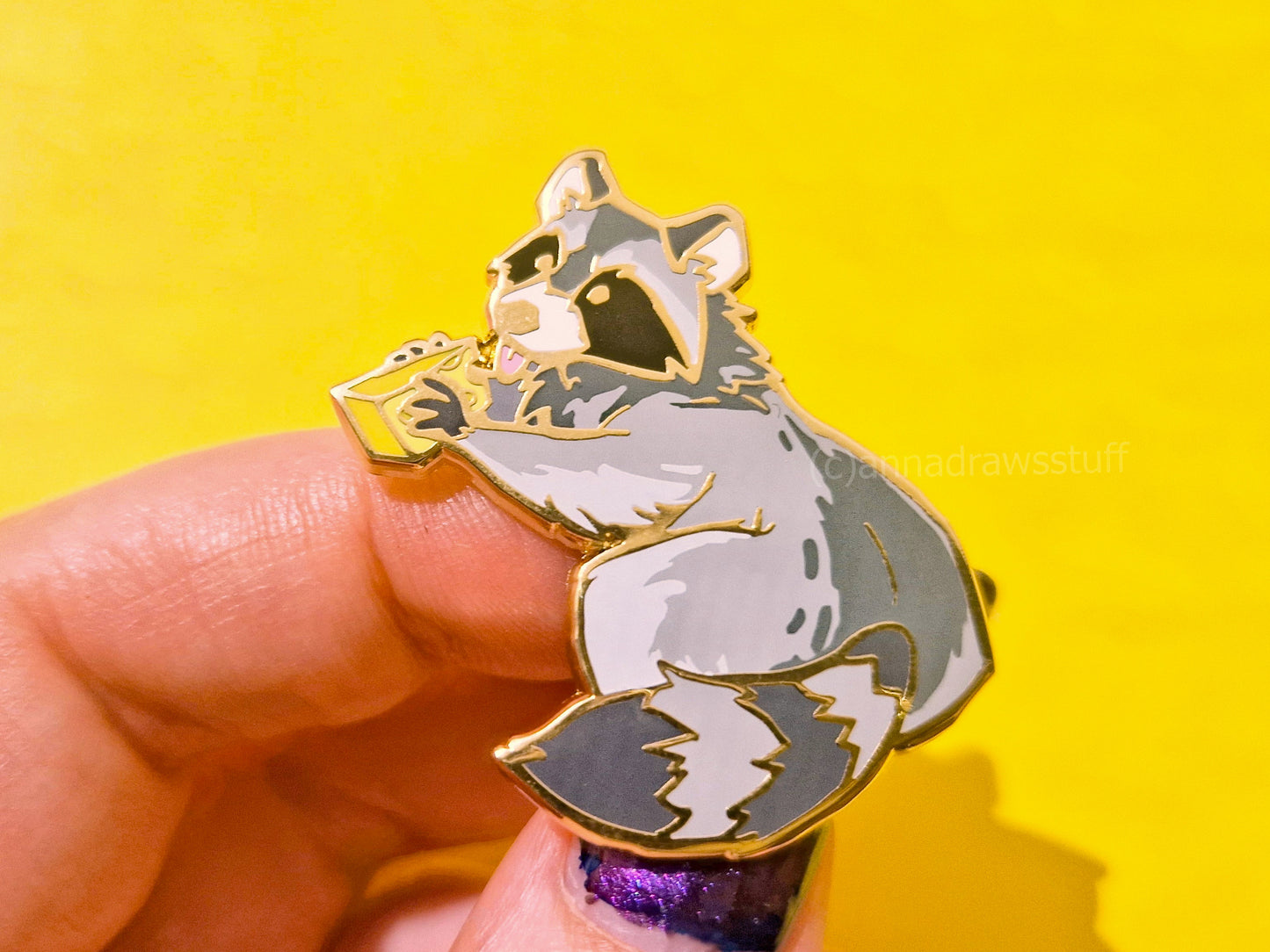 A hand holding an enamel pin of a raccoon holding a piece of cheese, set against a bright yellow background.