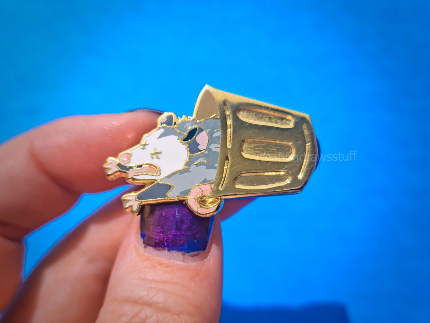 A hand with purple glitter nail polish holds a gold-tone enamel pin shaped like an opossum inside a trash can, set against a bright blue background.